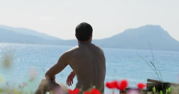 Pohledný muž sedí na mýtině před mořem, spousta květin máku, malebná mořská krajina při východu slunce, hory na pozadí, rozmazané sluneční odrazy na vodě — Stock video