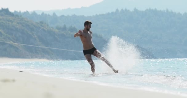 El hombre guapo con un cuerpo atlético perfecto en bañadores divirtiéndose en una playa desierta por la mañana, salpicando en el agua, jugando con las olas, mostrando su cuerpo y posando — Vídeos de Stock