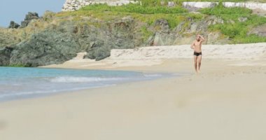 Yüzme şortu içinde mükemmel atletik vücudu olan yakışıklı bir adam sabahın köründe kumsalda eğleniyor, atlıyor ve el sallıyor, hızla sahilde koşuyor, vücudunu gösteriyor ve poz veriyor.