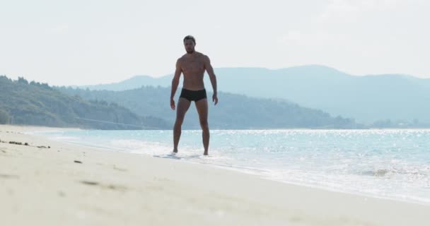 Ο όμορφος άνδρας με το τέλειο αθλητικό σώμα σε μαγιό διασκεδάζοντας σε μια ερημική παραλία το πρωί, καυχιέται για το σώμα ως bodybuilder, επιδεικνύοντας το σώμα του και ποζάροντας — Αρχείο Βίντεο