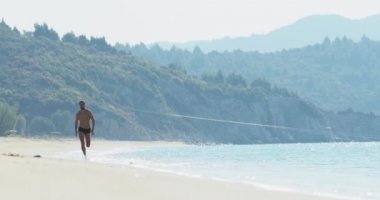Yüzme şortu içinde mükemmel atletik vücudu olan yakışıklı bir adam sabahın köründe ıssız bir plajda eğleniyor hızla koşuyor, vücudunu gösteriyor ve poz veriyor.