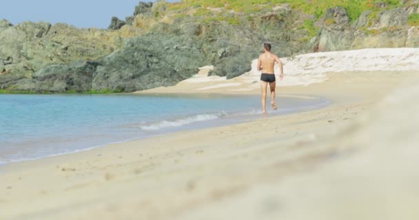 Pohledný muž s dokonalým atletickým tělem v plavkách se baví na opuštěné pláži v dopoledních hodinách, skáče jako vítěz, Rychle běží na pláži, předvádí své tělo a pózuje
