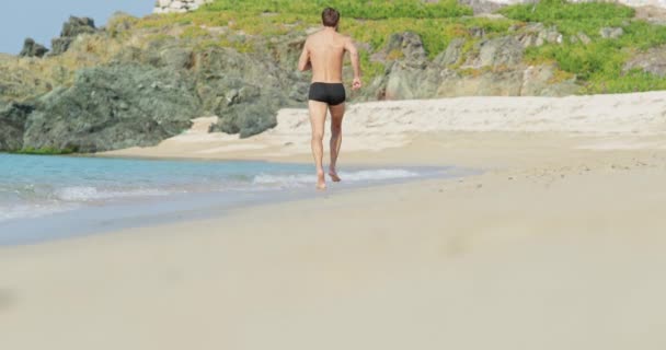 El hombre guapo con un cuerpo atlético perfecto corre lentamente en una playa alejándose de nosotros por la mañana, mostrando su cuerpo y posando, costa rocosa en el fondo — Vídeos de Stock