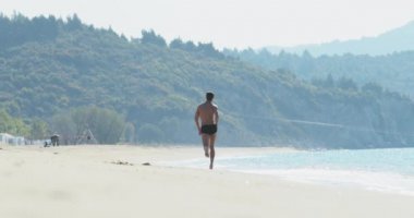 Yüzme şortu içinde harika bir vücudu olan yakışıklı bir adam sabahın köründe ıssız bir plajda eğleniyor, bir şampiyon gibi zıplıyor, hızla sahilde koşuyor, vücudunu gösteriyor ve poz veriyor.