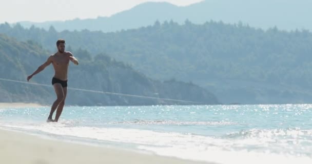 El hombre guapo con un cuerpo atlético perfecto en bañadores divirtiéndose en una playa desierta por la mañana, salpicando en el agua, jugando con las olas, mostrando su cuerpo y posando — Vídeos de Stock