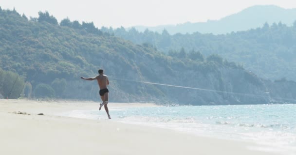 El hombre guapo con un cuerpo atlético perfecto en bañadores divirtiéndose en una playa desierta por la mañana, salpicando en el agua, jugando con las olas, mostrando su cuerpo y posando — Vídeos de Stock