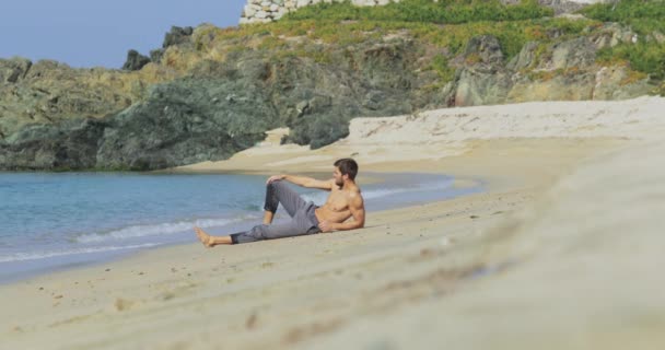 El hombre guapo con un cuerpo atlético perfecto en pantalones grises se sienta en una playa desierta por la mañana, mostrando su cuerpo y posando — Vídeos de Stock