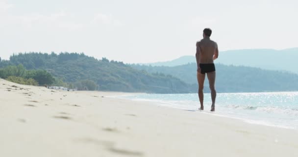 El hombre guapo con un cuerpo atlético perfecto en bañadores divirtiéndose en una playa desierta por la mañana, lentamente va como el macho, mostrando su cuerpo y posando — Vídeos de Stock
