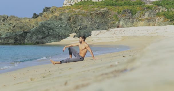 A jóképű férfi tökéletes atlétikai testtel, szürke nadrágban ül egy elhagyatott strandon reggel, mutogatja a testét és pózol. — Stock videók