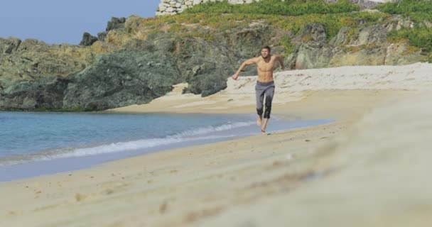 The handsome man with a perfect athletic body in gray trousers having fun on a deserted beach in the morning, He slowly runs on a beach, showing off his body and posing — Stock Video