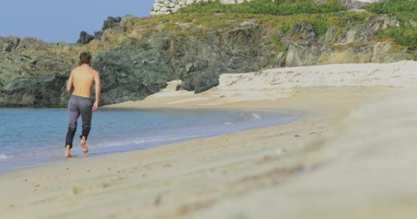 El hombre guapo con un cuerpo atlético perfecto en pantalones grises divirtiéndose en una playa desierta por la mañana, Corre lentamente en una playa, mostrando su cuerpo y posando — Vídeos de Stock