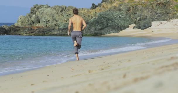 Ο όμορφος άνδρας με το τέλειο αθλητικό σώμα με γκρι παντελόνι που διασκεδάζει σε μια ερημική παραλία το πρωί, τρέχει αργά σε μια παραλία, επιδεικνύοντας το σώμα του και ποζάροντας — Αρχείο Βίντεο