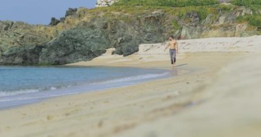 Gri pantolonlu, mükemmel atletik vücudu olan yakışıklı bir adam sabahın köründe kumsalda eğleniyor yavaşça kumsala gidiyor, vücudunu gösteriyor ve poz veriyor.