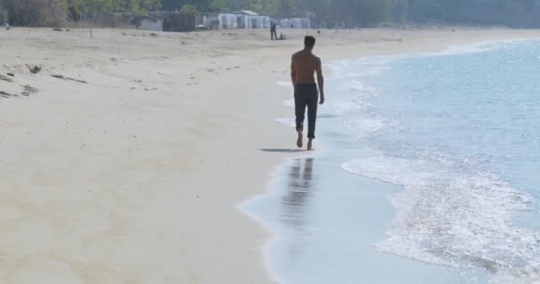 Ο όμορφος άνδρας με το τέλειο αθλητικό σώμα με γκρι παντελόνι που διασκεδάζει σε μια ερημική παραλία το πρωί, τρέχει αργά σε μια παραλία, επιδεικνύοντας το σώμα του και ποζάροντας — Αρχείο Βίντεο