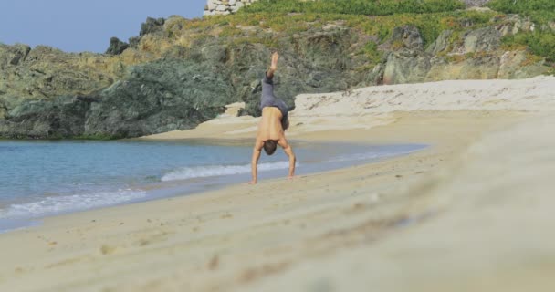 El hombre guapo con un cuerpo atlético perfecto en pantalones grises divirtiéndose en una playa desierta por la mañana, mostrando su cuerpo y posando — Vídeos de Stock