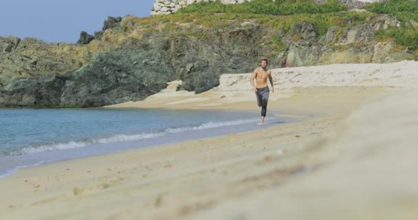 Przystojny mężczyzna z idealnym, wysportowanym ciałem w szarych spodniach bawiący się rano na bezludnej plaży, powoli biega po plaży, pokazując swoje ciało i pozując — Wideo stockowe