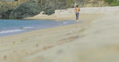Gri pantolonlu, mükemmel atletik vücudu olan yakışıklı bir adam, sabahın köründe kumsalda eğleniyor, yavaşça sahilde koşuyor, vücudunu gösteriyor ve poz veriyor.