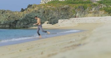 Gri pantolonlu, mükemmel atletik vücudu olan yakışıklı bir adam, sabahın köründe kumsalda eğleniyor, yavaşça sahilde koşuyor, vücudunu gösteriyor ve poz veriyor.
