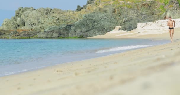 El hombre guapo con un cuerpo atlético perfecto en bañadores divirtiéndose en una playa desierta por la mañana, muy rápidamente corre por una playa, mostrando su cuerpo y posando — Vídeos de Stock