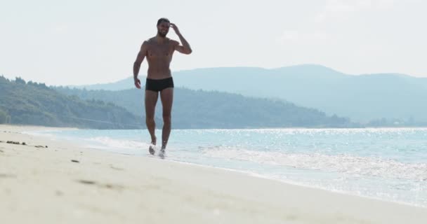 El hombre guapo con un cuerpo atlético perfecto en bañadores divirtiéndose en una playa desierta por la mañana, lentamente va como el macho, mostrando su cuerpo y posando — Vídeos de Stock