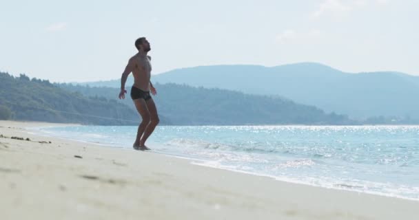 Przystojny mężczyzna z doskonałym atletycznym ciałem w kąpielówkach bawiący się rano na bezludnej plaży, skacze i macha rękami, rozgrzewa się przed bieganiem, pokazuje swoje ciało i pozuje — Wideo stockowe