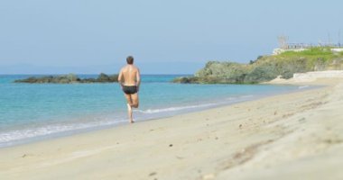 Yüzme şortu içinde harika bir vücudu olan yakışıklı bir adam sabahın köründe kumsalda eğleniyor yavaşça koşuyor, vücudunu gösteriyor ve poz veriyor.
