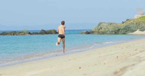 Ο όμορφος άνδρας με το τέλειο αθλητικό σώμα με μαγιό να διασκεδάζει σε μια ερημική παραλία το πρωί, τρέχει αργά σε μια παραλία, επιδεικνύοντας το σώμα του και ποζάροντας — Αρχείο Βίντεο