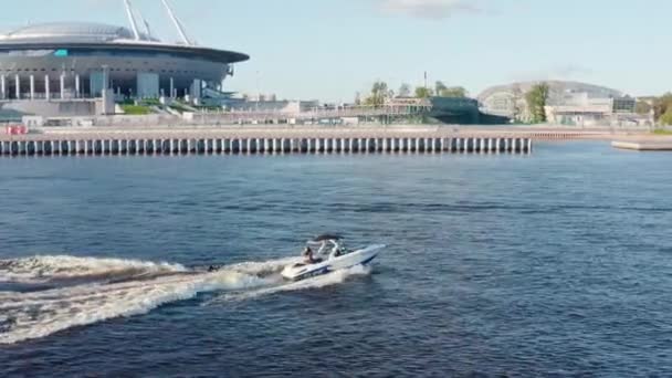 Rusya, St. Petersburg, 01 Eylül 2020: Gençler açık güneşli bir havada teknenin arkasında sörf tahtasına biniyor, Gazprom arenası ve kablo destekli bir köprü arka planda. — Stok video