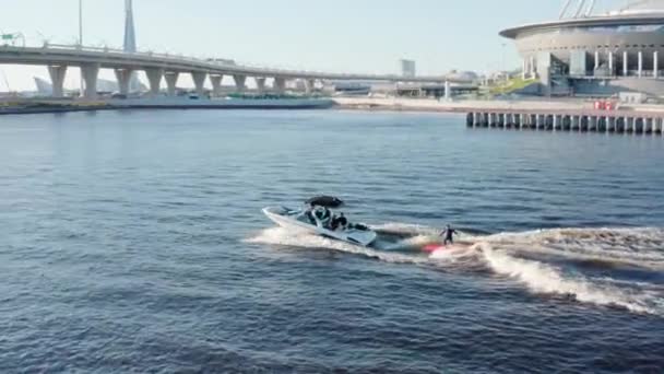 Rusland, St. Petersburg, 01 september 2020: Tieners rijden op een surfplank op het water achter de boot bij helder zonnig weer, de Gazprom arena en de kabelbrug op de achtergrond, wake surf — Stockvideo
