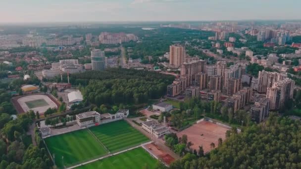 Ryssland, Sankt Petersburg, 18 juni 2021: Drönaren flyger över parken mot enorma bostadsområden under uppbyggnad vid solnedgången, Stadion för träningar av fotbollslaget Zenith, gröna träd — Stockvideo