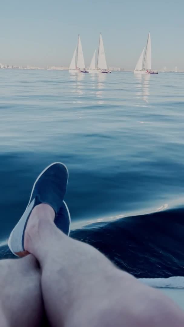 Ноги чоловіків одягнені в легке взуття лежать на краю човна, човен повільно плаває в ясну сонячну погоду, деякі вітрильники повільно йдуть на воду на фоні — стокове відео