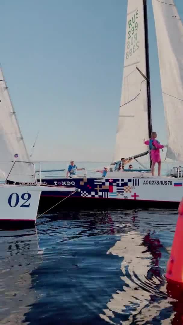 Ryssland, St Petersburg, 18 Juni 2021: Vissa segelbåtar i en lista går till sjöss, den klara himlen, regatta, Gazprom arena stadion på bakgrunden, reflektion av segel på vattnet, bro på bakgrunden — Stockvideo