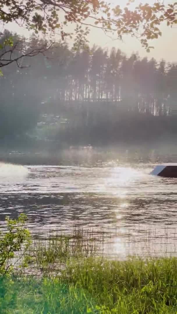 Vertikal video av en sjö vid solnedgången, semesterfirare simma på en uppblåsbar bräda och paddla, killen har roligt ridning styrelsen för repet, hoppa från språngbräda, wakeboard, stänk vatten — Stockvideo