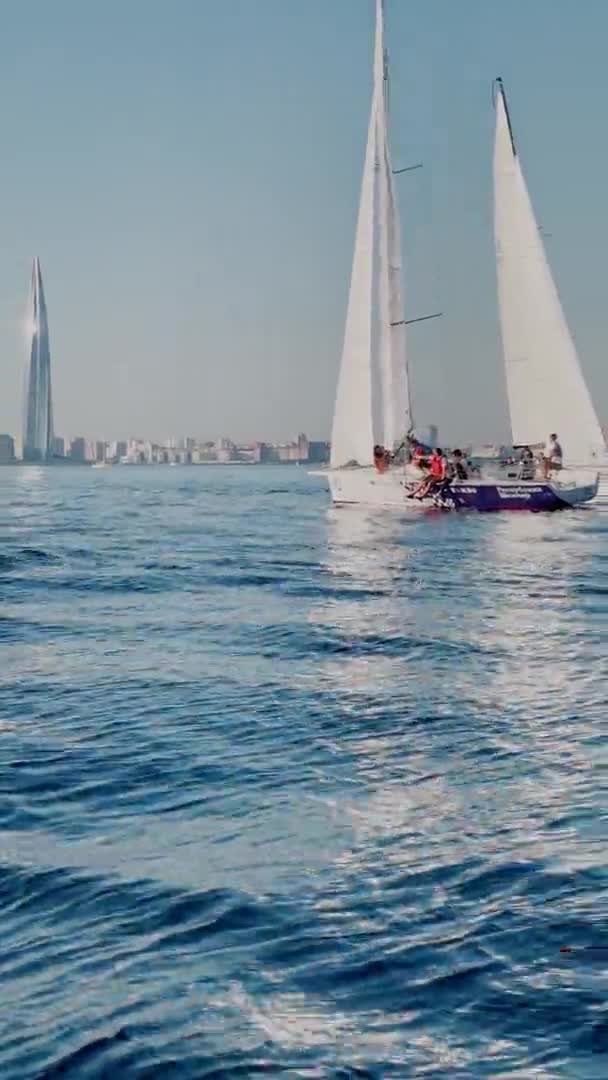 Russia, St.Petersburg, 18 June 2021: Some sailboats in a list goes by sea, the clear sky, regatta, Gazprom arena stadium on background, reflection of sail on the water, bridge on background — Stock Video