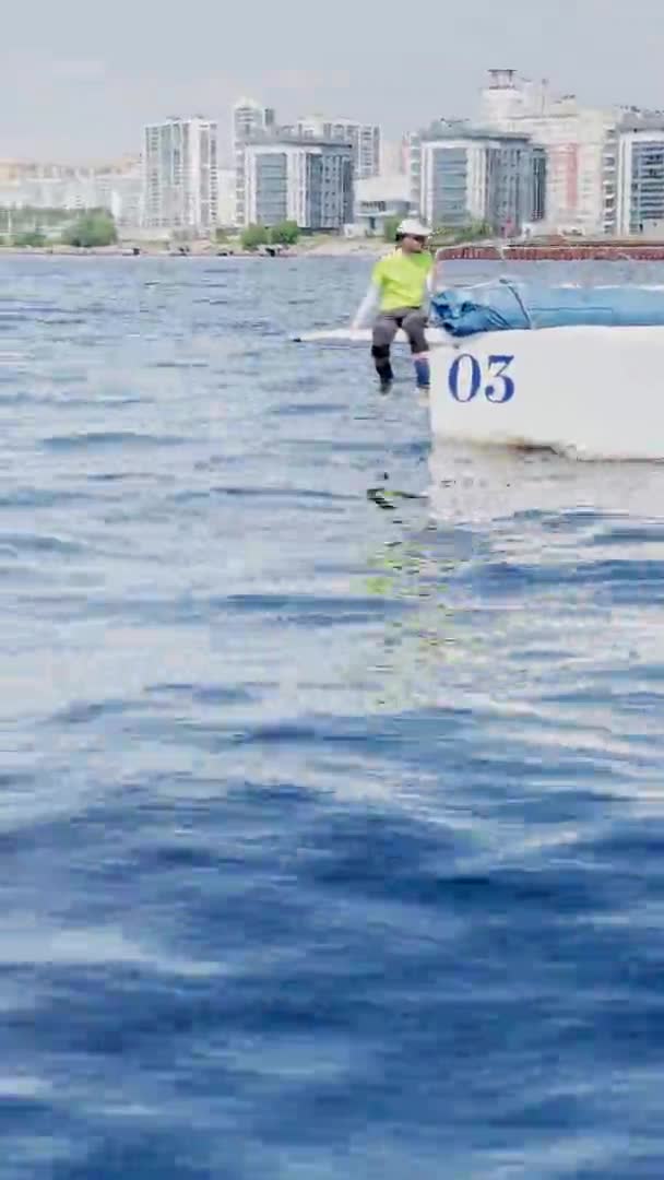 Russia, San Pietroburgo, 20 giugno 2021: La barca a vela va ad alta velocità con il tempo limpido e soleggiato, lo yachtsman in costume e occhiali da sole siede sul bompresso con le gambe pendenti — Video Stock