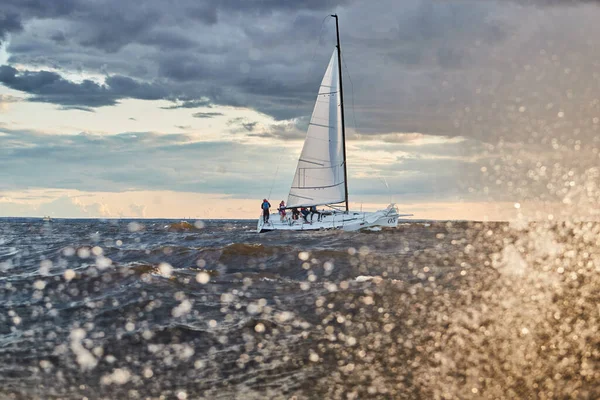 Das einsame Segelboot am Horizont durch Spritzer Meer bei Sonnenuntergang, der Gewitterhimmel in verschiedenen Farben, große Wellen, Segelregatta, bewölktes Wetter, nur Großsegel, Sonnenstrahlen — Stockfoto