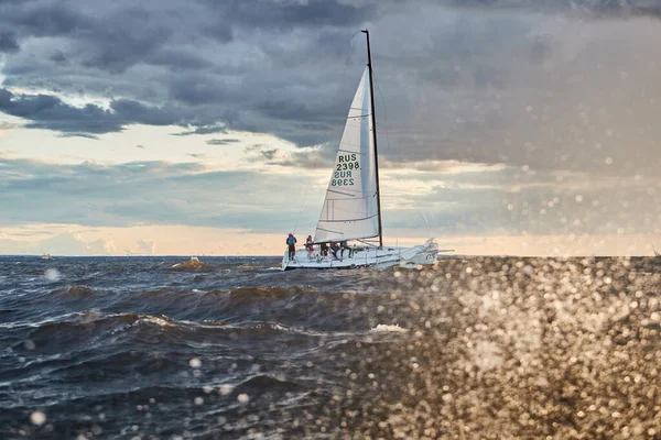 Russland, St. Petersburg, 23. Juli 2021: Das einsame Segelboot am Horizont durch Meeresplätschern bei Sonnenuntergang, der Gewitterhimmel in verschiedenen Farben, große Wellen, Segelregatta, bewölktes Wetter, Sonnenstrahlen — Stockfoto