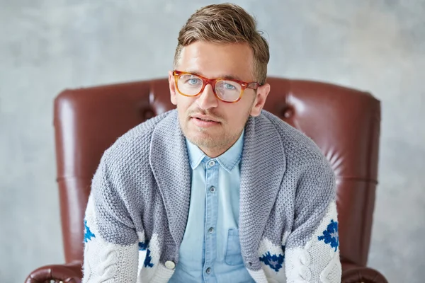 Retrato de un joven hombre inteligente mira a la cámara, buena vista, pequeño sin afeitar, carismático, con gafas llenas de marrón, camisa azul, suéter gris, sentado en una silla de cuero marrón sobre un fondo gris —  Fotos de Stock