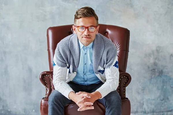 Retrato de un joven hombre inteligente mira a la cámara, buena vista, pequeño sin afeitar, carismático, con gafas llenas de marrón, camisa azul, suéter gris, sentado en una silla de cuero marrón sobre un fondo gris — Foto de Stock