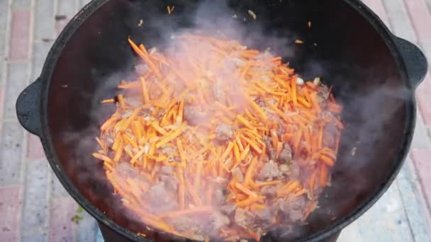 Pilaf plato nacional uzbeko en un gran caldero de hierro fundido en el fuego, añadir zanahorias rojas en rodajas, arroz, cordero, grasa de cordero, la cola de grasa, aceite de sésamo, cebolla, ajo, chile rojo picante — Vídeos de Stock