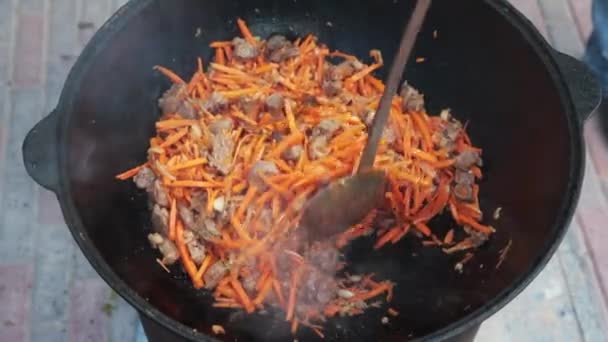 Usbekisches Nationalgericht pilaf in einem großen gusseisernen Kessel auf dem Feuer, fügen Sie in Scheiben geschnittene rote Karotten, Reis, Lamm, Hammelfett, den fetten Schwanz, Sesamöl, Zwiebeln, Knoblauch, rote Chilischote — Stockvideo