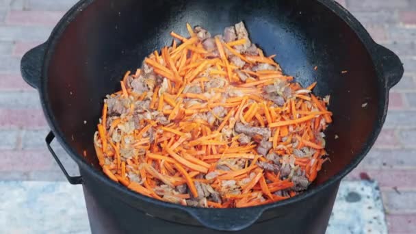 Pilaf masakan nasional Uzbekistan dalam kuali besi cor besar di atas api, tambahkan irisan wortel merah, beras, domba, lemak kambing, ekor lemak, minyak wijen, bawang, bawang putih, cabai merah — Stok Video