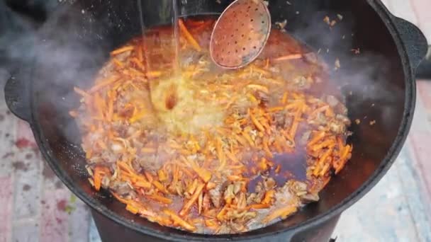 Uzbek national dish pilaf in a large cast-iron cauldron on the fire, add sliced red carrots, pour cold water, rice, lamb, mutton fat, the fat tail, sesame oil, onion, garlic, red hot chili pepper — Stock Video