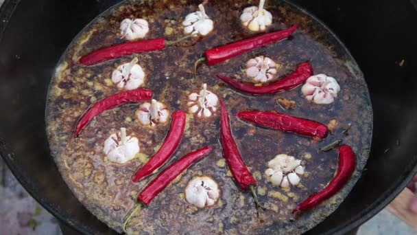 Uzbek national dish pilaf in a large cast-iron cauldron on the fire, sliced red carrots, cold water, add whole of garlic and red chili peppers, rice, lamb, mutton fat, the fat tail, sesame oil, onion — Stock Video