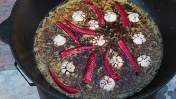 Pilaf plato nacional uzbeko en un gran caldero de hierro fundido en el fuego, zanahorias rojas en rodajas, agua fría, añadir todo el ajo y chiles rojos, arroz, cordero, grasa de cordero, la cola de grasa, aceite de sésamo, cebolla — Vídeos de Stock