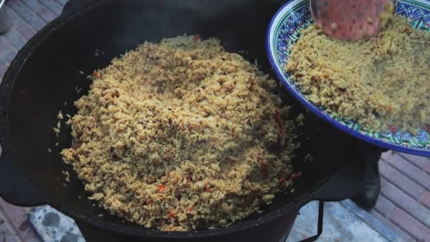 Uzbecká národní miska pilaf ve velkém litinovém kotlíku na ohni, roztáhl pilaf na velkém talíři, mrkev, Barberry, česnekový tuk, papriku z chilli, rýži, jehněčí, skopové maso, tlustý ocas, sezamový olej — Stock video