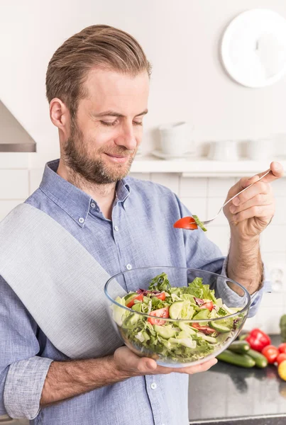 Feliz hombre sonriente comiendo ensalada de verduras frescas en la cocina — Foto de Stock