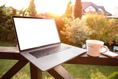 Laptop bilgisayar (boş ekranlı) ve bahçe terasında kahve fincanı. Günaydın. Arka planda doğan güneş..