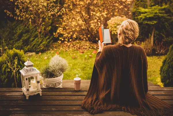Blond woman reading book outdoor on the garden terrace. Autumn (fall) garden as background. Countryside or suburb lifestyle - leisure.