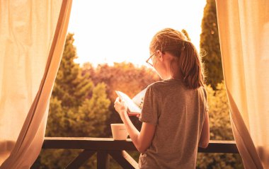 Beyaz çocuk bahçedeki terasta kitap okuyor. Çocukluk ve eğitim. Altın gün batımı ışığı. Kırsal ya da banliyö yaşam tarzı. 